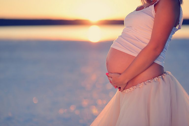 Image of pregnant woman cradling her baby bump on the beach. The sun is setting in the background.